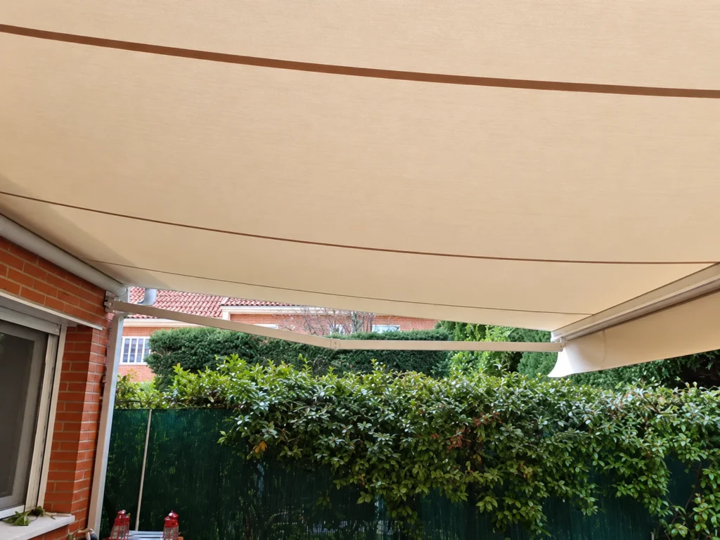 Toldos para balcones en Madrid: toldo extensible de lona beige junto a una ventana y un seto